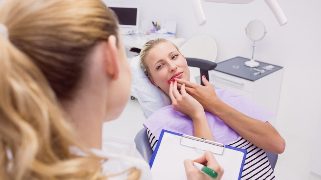 Patient describing tooth sensitivity to a dentist A blonde woman in a dental chair pointing to her jaw and discussing tooth pain or sensitivity with a dentist during a consultation.